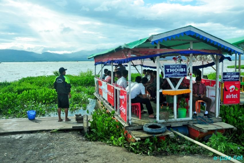 Vibrant floating café scene at Loktak Lake. Floating lakeside café with locals gathered under a green roof overlooking calm waters.
